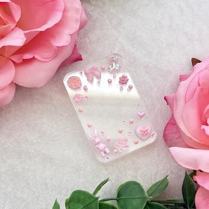 May include: Clear rectangular phone case decorated with pink bows, hearts, stars, a bunny, and a teddy bear. The case is surrounded by pink roses and green leaves.
