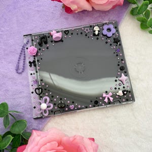 May include: A square clear plastic CD case decorated with various small charms including a pink rubber duck, purple flowers, black hearts, a bow, and small beads in shades of black and purple. A purple beaded chain is attached to the side of the case.
