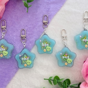 May include: Five star-shaped keychains with silver clasps. Each keychain is light blue with glitter and small decorative elements inside. The keychains have different text: "atz", "skz", "bts", "svt", and "en-".