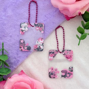 May include: Two clear acrylic keychains with pink and black cartoon character designs and attached burgundy ball chains. The keychains have a square shape with rounded corners and are decorated with various cute illustrations.