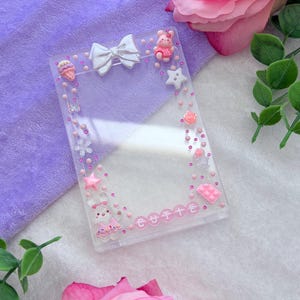 May include: A clear rectangular case decorated with pink and white embellishments. The case features a white bow, a pink bear, stars, and the word "CUTTE". The case is surrounded by pink dots and small flowers.
