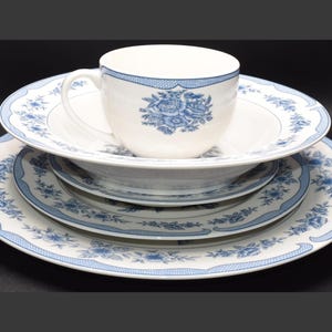 May include: A white porcelain teacup and saucer set with a blue floral design. The set includes a teacup, a saucer, a bowl, and two plates. The plates and teacup have a blue floral border.