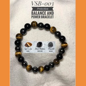Può includere: Un braccialetto realizzato con perline di ematite nera e occhio di tigre. Il braccialetto è etichettato "VSB-003 Protection BALANCE AND POWER BRACELET". Le perline sono disposte in un motivo nero e oro. Il braccialetto è anche etichettato con i nomi delle pietre e le loro proprietà: "Occhio di Tigre - Potere", "Obsidiana Nera - Protezione" e "Ematite - Equilibrio".