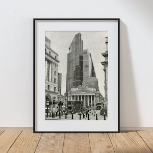 May include: Black and white framed print of a city street scene. The image features tall buildings, including skyscrapers and classical architecture, with a double-decker bus and pedestrians. The print is displayed on a wooden floor against a white wall.