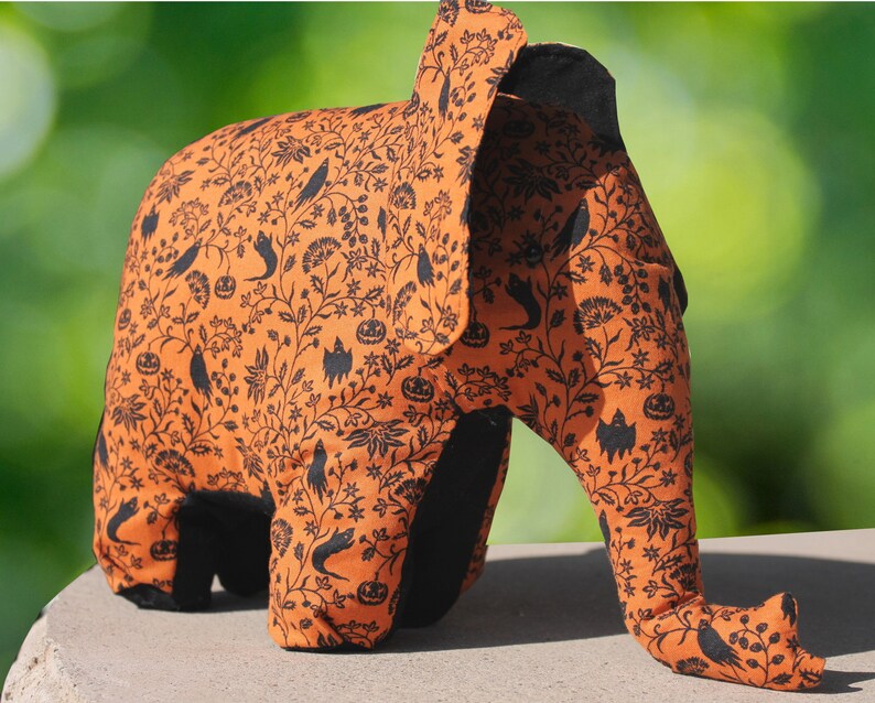 May include: Orange and black Halloween elephant plush toy with a black and white floral pattern. The elephant has a long trunk and large ears.