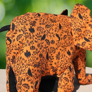 May include: Orange and black fabric elephant with a Halloween-themed print. The print features black bats, pumpkins, and other Halloween symbols.