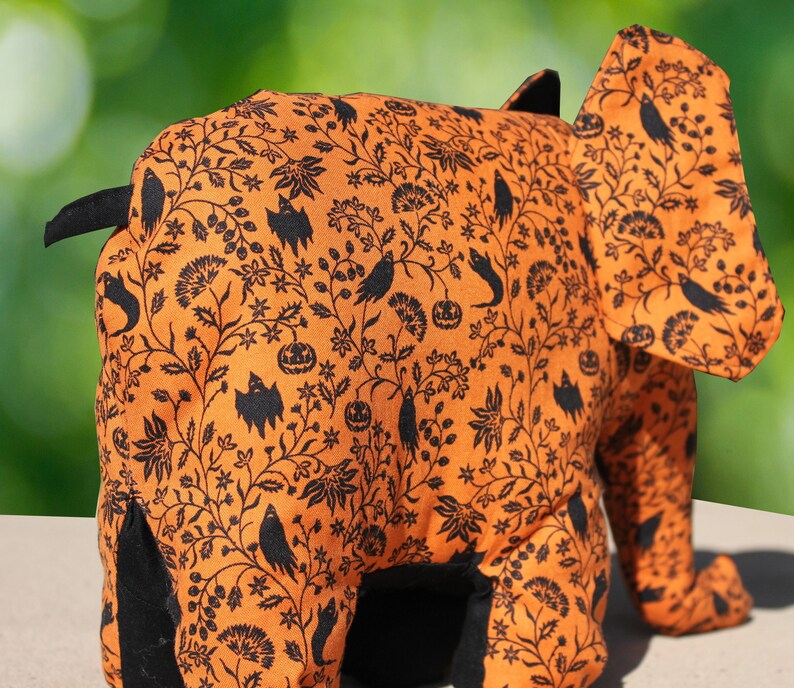 May include: Orange and black fabric elephant with a Halloween-themed print. The print features black bats, pumpkins, and other Halloween symbols.