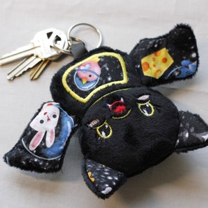 May include: Black plush keychain with a space-themed design. The keychain has a round head with embroidered eyes and a red nose. The keychain has two wings with different space-themed patterns, including a bunny, a planet, and a cheese.