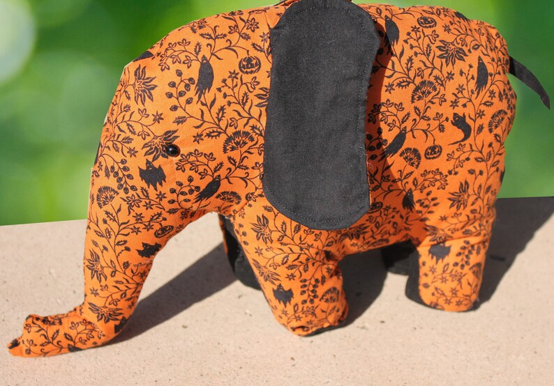 May include: Orange and black fabric elephant with a Halloween-themed print. The print features black bats, birds, and pumpkins on a repeating pattern.
