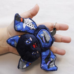 This bat plushie is 4 1/2' tall and 6 1/2' wide from wing to wing. It is made from dark blue minky fabric and a cotton fabric with sharks in Halloween costumes on it for the ears, wings, and belly. The belly is outlined in blue thread as well as the eyes. It has small white fangs and pink cheeks embroidered on its face. As well as a red mouth and red heart shaped nose.
The keychain is as big as the hand holding it.

Keys not included.