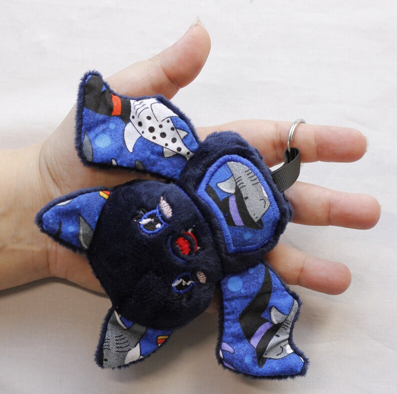 This bat plushie is 4 1/2' tall and 6 1/2' wide from wing to wing. It is made from dark blue minky fabric and a cotton fabric with sharks in Halloween costumes on it for the ears, wings, and belly. The belly is outlined in blue thread as well as the eyes. It has small white fangs and pink cheeks embroidered on its face. As well as a red mouth and red heart shaped nose.
The keychain is as big as the hand holding it.

Keys not included.