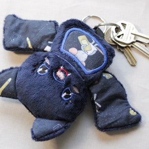 May include: A blue plush toy shaped like a monster with a key chain attached. The toy has a small pocket on its chest with an embroidered image of a cartoon character. The toy has a black eye and a red mouth.