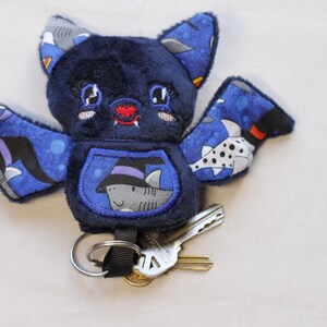 Unedited Photo: This bat plushie is 4 1/2' tall and 6 1/2' wide from wing to wing. It is made from dark blue minky fabric and a cotton fabric with sharks in Halloween costumes on it for the ears, wings, and belly. The belly is outlined in blue thread as well as the eyes. It has small white fangs and pink cheeks embroidered on its face. As well as a red mouth and red heart shaped nose.

Keys not included.