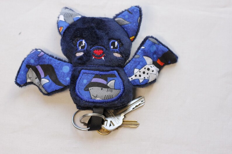 Unedited Photo: This bat plushie is 4 1/2' tall and 6 1/2' wide from wing to wing. It is made from dark blue minky fabric and a cotton fabric with sharks in Halloween costumes on it for the ears, wings, and belly. The belly is outlined in blue thread as well as the eyes. It has small white fangs and pink cheeks embroidered on its face. As well as a red mouth and red heart shaped nose.

Keys not included.