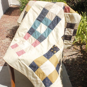 May include: A cream-colored quilt with a patchwork design featuring squares of various colors and patterns, including blue, green, red, and yellow. The quilt is folded and draped over a wooden chair.
