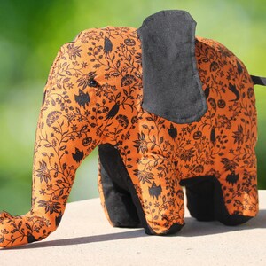 May include: A plush orange and black elephant toy with a Halloween-themed print. The elephant has a black fabric ear and a black fabric tail.