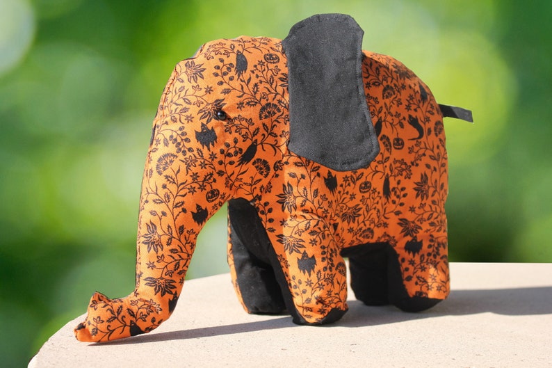 May include: A plush orange and black elephant toy with a Halloween-themed print. The elephant has a black fabric ear and a black fabric tail.