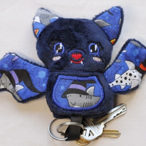 This bat plushie is 4 1/2' tall and 6 1/2' wide from wing to wing. It is made from dark blue minky fabric and a cotton fabric with sharks in Halloween costumes on it for the ears, wings, and belly. The belly is outlined in blue thread as well as the eyes. It has small white fangs and pink cheeks embroidered on its face. As well as a red mouth and red heart shaped nose.

Keys not included.