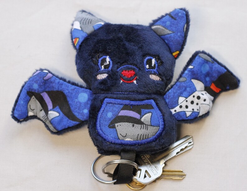 This bat plushie is 4 1/2' tall and 6 1/2' wide from wing to wing. It is made from dark blue minky fabric and a cotton fabric with sharks in Halloween costumes on it for the ears, wings, and belly. The belly is outlined in blue thread as well as the eyes. It has small white fangs and pink cheeks embroidered on its face. As well as a red mouth and red heart shaped nose.

Keys not included.