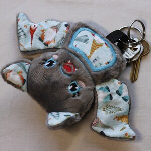 May include: Gray plush bat keychain with blue and white fabric ears featuring a whimsical pattern of fish and trees. The keychain has a silver key ring and a black strap.