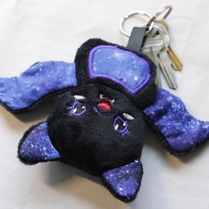 May include: A plush black bat keychain with a galaxy print on its wings. The bat has a purple outline and red embroidered eyes. The keychain is attached to a silver key ring.