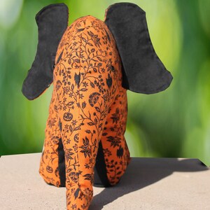 May include: A plush orange and black elephant with a floral pattern. The elephant is sitting on a light brown surface.