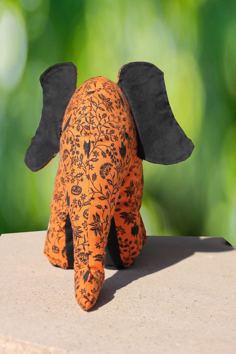 May include: A plush orange and black elephant with a floral pattern. The elephant is sitting on a light brown surface.