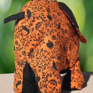 May include: Orange and black Halloween-themed fabric elephant with a black floral pattern. The elephant is standing on a light brown surface.
