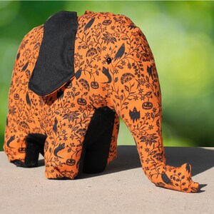 May include: A plush orange and black elephant toy with a Halloween-themed print. The print features black bats, pumpkins, and other Halloween imagery on a light orange background.