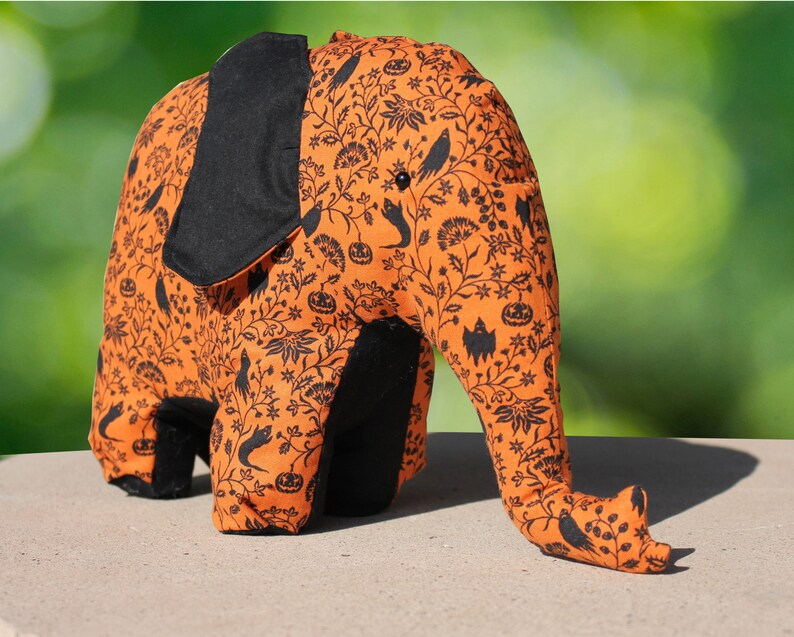 May include: A plush orange and black elephant toy with a Halloween-themed print. The print features black bats, pumpkins, and other Halloween imagery on a light orange background.