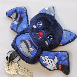 This bat plushie is 4 1/2' tall and 6 1/2' wide from wing to wing. It is made from dark blue minky fabric and a cotton fabric with sharks in Halloween costumes on it for the ears, wings, and belly. The belly is outlined in blue thread as well as the eyes. It has small white fangs and pink cheeks embroidered on its face. As well as a red mouth and red heart shaped nose.

Keys not included.