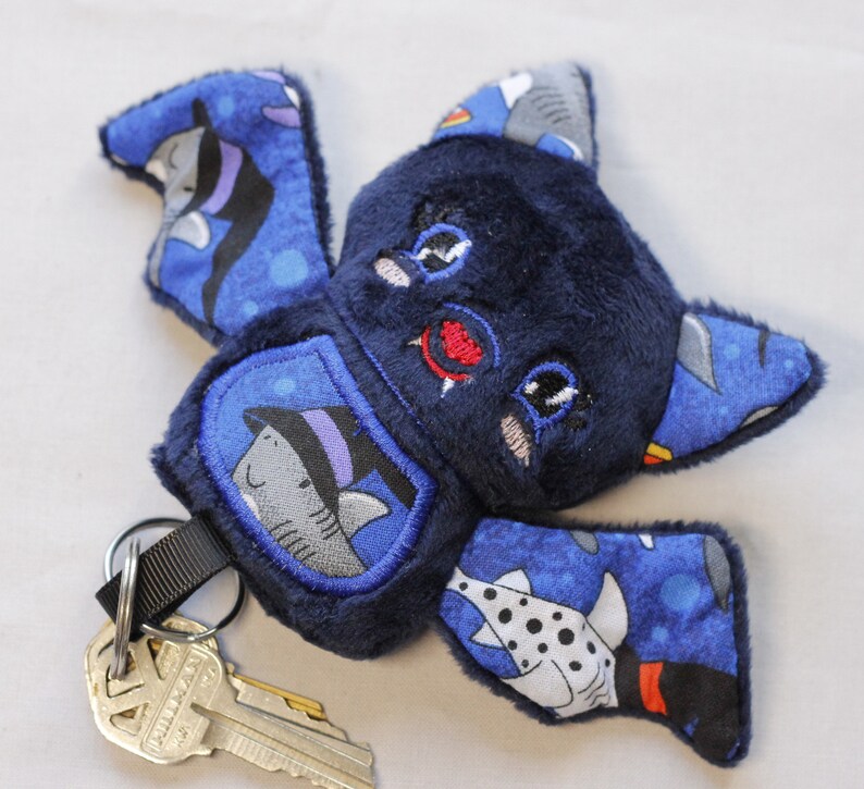 This bat plushie is 4 1/2' tall and 6 1/2' wide from wing to wing. It is made from dark blue minky fabric and a cotton fabric with sharks in Halloween costumes on it for the ears, wings, and belly. The belly is outlined in blue thread as well as the eyes. It has small white fangs and pink cheeks embroidered on its face. As well as a red mouth and red heart shaped nose.

Keys not included.