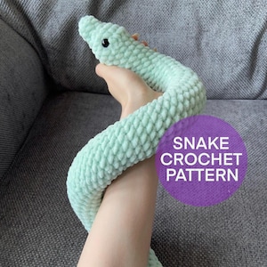 Puede incluir: Un juguete de serpiente de crochet de color verde claro. La serpiente está enroscada en la mano de una persona. El texto "SNAKE CROCHET PATTERN" está en un círculo morado en el fondo.
