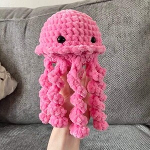 May include: A handmade pink crochet jellyfish toy with black button eyes and long, curly tentacles. The plush toy is made of soft, textured yarn, and is held against a gray background.