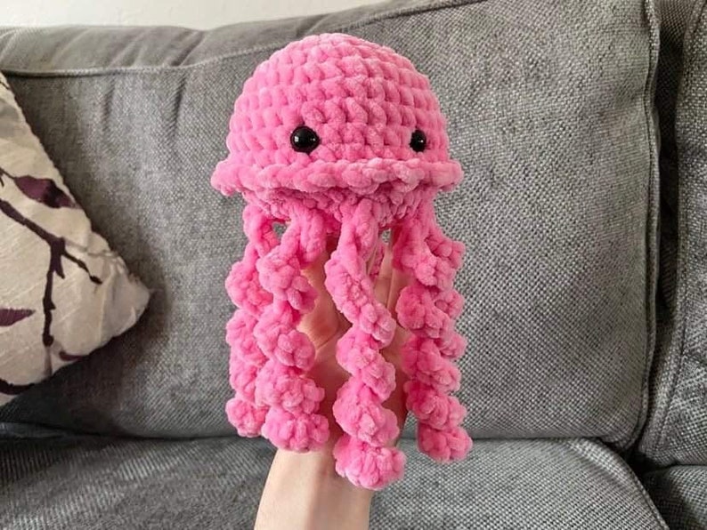May include: A handmade pink crochet jellyfish toy with black button eyes and long, curly tentacles. The plush toy is made of soft, textured yarn, and is held against a gray background.