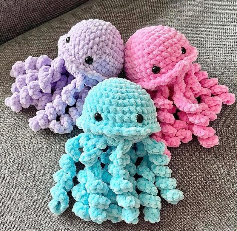 May include: Three handmade crochet jellyfish in shades of purple, pink, and blue. Each jellyfish has a rounded head with black button eyes and long, curly tentacles. The soft, plush texture and vibrant colors make them appealing.