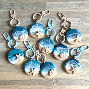 May include: A selection of ocean-themed keyrings. Each keyring has a round design depicting a beach scene with a blue wave pattern, a beige sand base, and a seashell or starfish. They have metal clasps.