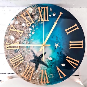 May include: A round blue and white resin clock with gold Roman numeral markings and gold hands. The clock face features a design of sand, seashells, and a starfish.
