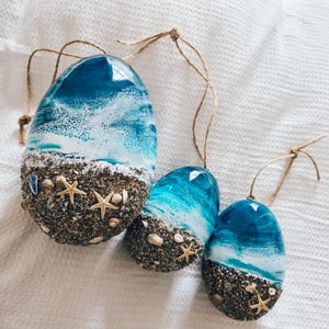 May include: Three resin ornaments with a beach theme. Each has a glossy blue and white wave design at the top, and a textured base of sand, shells, and starfish. They are suspended by twine loops. The ornaments are ideal for coastal decor.