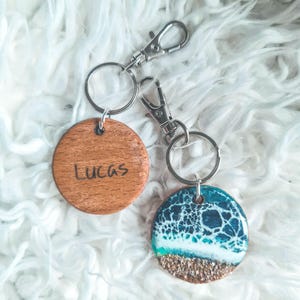 May include: Two keychains with round wooden and resin charms. The wooden charm is engraved with the name "Lucas". The resin charm features a blue and white ocean wave design with a sandy bottom.