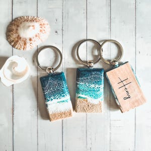 May include: Three keychains with ocean-themed resin charms. Two charms feature blue and white resin with a sandy bottom. The third charm is a wooden rectangle with the name "Henry" written on it.