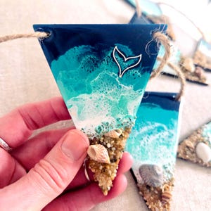 May include: Triangular beach-themed ornament with a dark blue top, transitioning to turquoise and white, resembling ocean waves. A silver whale tail charm is attached. The bottom edge is adorned with sand and seashells, and a jute string is attached for hanging.