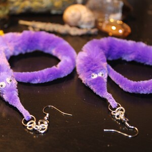Può includere: Un paio di orecchini a forma di verme peloso viola con occhietti. Gli orecchini sono attaccati a ganci d'argento.