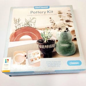 Peut inclure: Une boîte de kit de poterie Craftmaker de conception classique. La boîte présente une pièce de poterie en forme d'arche de couleur terre cuite, un petit pot brun foncé avec une succulente et un vase vert avec de l'eucalyptus. Le kit comprend un livre de 24 pages et de l'argile.