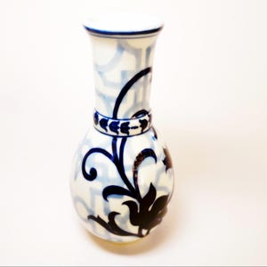 May include: A white ceramic vase with blue and black floral and geometric patterns. The vase has a rounded base, a narrow neck, and a flared rim. A dark blue band encircles the neck. The vase is likely for decorative purposes.