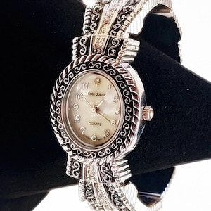 Può includere: Un orologio al quarzo color argento con quadrante bianco e dettagli ornati. L'orologio presenta un quadrante rotondo con numeri romani e una lunetta decorativa. Il cinturino ha un design intricato con piccoli abbellimenti. L'orologio è marchiato "Cote d'Azur" e "Japan Movt".