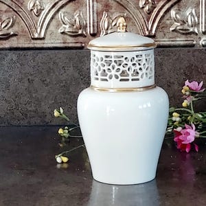 May include: A white ceramic jar with a lid, featuring a gold trim and a decorative openwork design. The jar has a glossy finish and a gold finial on the lid. The background includes a textured gold panel and a dark surface.