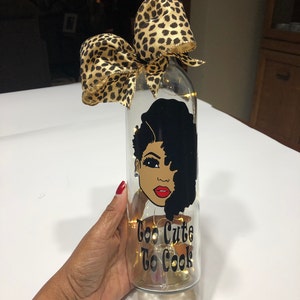 May include: A clear glass bottle with a cork top and a leopard print ribbon. The bottle has a black and white illustration of a woman with an afro and the text "Too Cute To Cook".