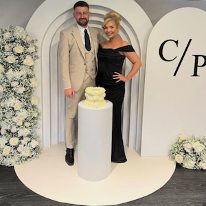 May include: A couple pose for a wedding photo in front of a white archway with the letters "C/P" on the right side. The couple are standing in front of a white cake on a white pedestal. The woman is wearing a black dress and the man is wearing a tan suit.