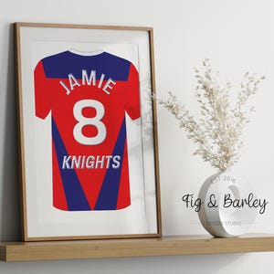 May include: A framed print of a red, blue, and white jersey with the name "Jamie" and the number "8" on the back. The jersey also has the word "Knights" printed on the back. The frame is brown wood.